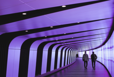 Two men walk away from the camera as they stroll through a modern tunnel with glass on one side and stripes on the other.