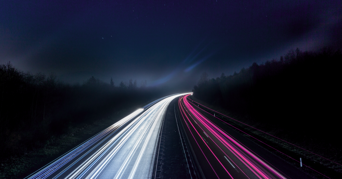 Stylized stop motion photo of a dark forest transected by a highway with streaks of light depicted as head and tail lights.