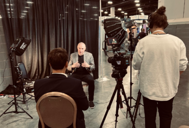 A man gestures with his hands as he speaks while being interviewed by a reporter. A camera operator is also shown.